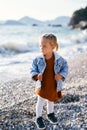 Little pensive girl stands on a pebble beach, holding onto the rim of a denim jacket and looks into the distance Royalty Free Stock Photo