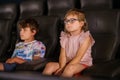 Little preschool girl with glasses and kid boy watching cartoon movie in cinema and eating popcorn. Happy excited Royalty Free Stock Photo