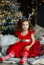 Little pretty curly smiling girl sitting nearly Christmas tree with Christmas decorations and presents. Child in red dress and soc