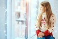 A little pretty sad blonde girl in a winter sweater sits on the window and looks into the distance. quarantine. Royalty Free Stock Photo