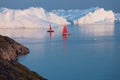 Little red sailboat cruising among floating icebergs in Disko Bay glacier during midnight sun season of polar summer. Greenland