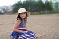 Little sad girl sitting in summer hat and beautiful dress on the beach Royalty Free Stock Photo