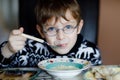 Little school boy with glasses eating vegetable soup indoor. Blond child in domestic kitchen or in school canteen. Cute Royalty Free Stock Photo