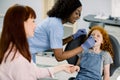 Little school girl with red curly hair, visiting dentist for checkup or caries treatment, sitting in dental chair and