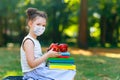 Little school kid girl wearing medical mask. Child holding different colorful books, glasses and apple. Closed schools
