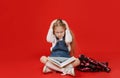 a little schoolgirl girl is sitting with a book upset with her hands on her head, worrying on a red background