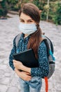 Little schoolgirl with a medical mask on her face and a laptop in her hand goes to school