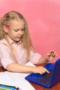 little schoolgirl is typing on a laptop at the table. pink background.