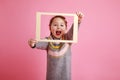 Little screaming girl in warm dress looking through frame on rosa bachground