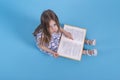 A  little serious girl  reads book on blue background with copy space in studio, top view Royalty Free Stock Photo