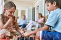 Little siblings playing chess at porch with parents sitting in background