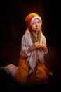 Little skinny girl in long white and yellow dress with scarf on head. Young model posing for photo shoot in dark studio
