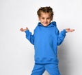 Little sly girl in a blue sports suit shrugs while standing on a white background.