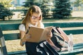 Little smart girl in glasses with a school backpack reading book sitting on bench in the park Royalty Free Stock Photo