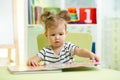 Little smart girl looking at book while sitting on chair in nursery Royalty Free Stock Photo