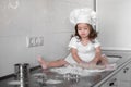 Little smiling baby girl baker in white cook hat and apron kneads a dough on tle kitchen Royalty Free Stock Photo