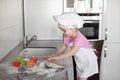 Little smiling baby girl baker in white cook hat and apron kneads a dough on tle kitchen