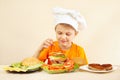 Little smiling boy in chefs hat puts tomato on hamburger Royalty Free Stock Photo