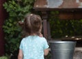 Little smiling country girl looks thoughtfully at the metal bucket on the well