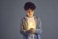 Little smiling curly boy stands on a gray background and looks at a bright light bulb in his hands. Royalty Free Stock Photo