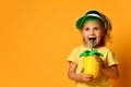Little smiling cute blond girl in yellow t-shirt and hat drinking fresh healthy fruit juice from straw over yellow background Royalty Free Stock Photo