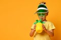 Little smiling cute blond girl in yellow t-shirt and hat drinking fresh healthy fruit juice from straw over yellow background Royalty Free Stock Photo