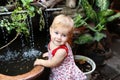 Little smiling girl with blond hair and blue eyes in sundress is standing in garden with pot plants and waterfall Royalty Free Stock Photo