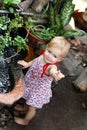 Little smiling girl with blond hair and blue eyes in sundress is standing in garden with pot plants and waterfall Royalty Free Stock Photo