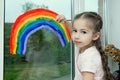 Little smiling girl sits by the window on the windowsill and draws a rainbow on the glass Royalty Free Stock Photo