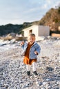 Little smiling girl in a dress and denim jacket walks along the pebble beach against the backdrop of the sea, rocks and Royalty Free Stock Photo