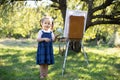 Little smiling girl in jeans dress, painting at green summer park outdoor. Talented small girl artist, holding brush in Royalty Free Stock Photo