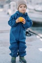 Little smiling girl in overalls with ripe persimmon stands on the pier Royalty Free Stock Photo