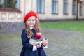Little smiling girl in a red beret holds a stack of books with an apple near school building. Cute schoolgirl of primary school on Royalty Free Stock Photo