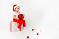 Little smiling girl in santa helper hat sitting on a giftbox with red ribbon and holding red christmas ball, white isolated Royalty Free Stock Photo