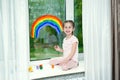 Little smiling girl sits by the window on the windowsill and draws a rainbow on the glass Royalty Free Stock Photo