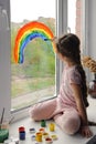 Little smiling girl sits by the window on the windowsill and draws a rainbow on the glass Royalty Free Stock Photo