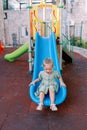 Little smiling girl sitting at the bottom of a plastic slide on a playground in the yard