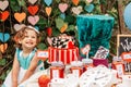 Little smiling girl dressed as Alice in Wonderland near table with fairy tale props. Cake, Hatter hat, clock, card suits Royalty Free Stock Photo