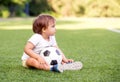 Little soccer fan sitting at football field with ball, looking at stadium. Side view. Toddler boy watching game with serious face Royalty Free Stock Photo