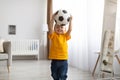Little sportsman. Adorable little boy posing with football ball, lifting it up in hands, standing in living room