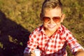 Little stylish boy in sunglasses is sitting on the meadow drinki