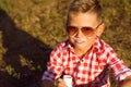 Little stylish boy in sunglasses is sitting on the meadow drinking yogurt