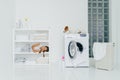 Little tired girl did work about house to help parents, sleeps on shelf near folded linen, iron and washing powder containers,