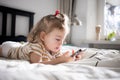 Little toddle girl playing with smartphone laying on the bed at home. Caucasian toddler girl.