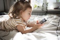 Little toddle girl playing with smartphone laying on the bed at home. Caucasian toddler girl.