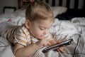 Little toddle girl playing with smartphone laying on the bed at home. Caucasian toddler girl.