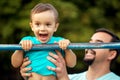 Little toddler boy doing pull ups exercise, child smiling happily having pulled up on shabby bar with support of father.