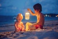 Little toddler boy, sitting on the beach after sunset on a moonlight with a lantern, enjoying the quiteness of the beach