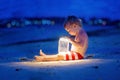 Little toddler boy, sitting on the beach after sunset on a moonlight with a lantern, enjoying the quiteness of the beach