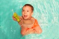 Little toddler boy sitting in swimming pool with funny face expression and shooting or aiming up with green water gun Royalty Free Stock Photo